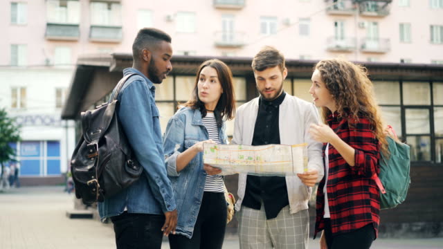Multiracial-group-of-tourists-is-looking-at-paper-map-standing-in-the-street-in-modern-city-talking-and-laughing.-Travelling,-navigation-and-youth-concept.
