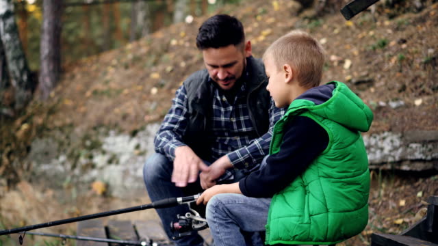 Bearded-young-man-is-fishing-with-cute-child-on-autumn-day,-boy-is-holding-rod-and-talking-to-father-learning-to-catch-fish.-People,-sharing-experience-and-family-concept.