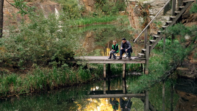 Loving-father-is-teaching-his-son-to-catch-fish-on-autumn-day,-people-are-sitting-on-wooden-pier-with-rods-and-talking-sharing-experience.-Recreation-and-family-concept.