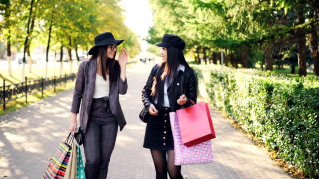 Two-attractive-young-women-are-walking-from-shops-along-beautiful-street-carrying-paper-bags-with-purchases-and-talking-on-beautiful-autumn-day.-Shopping-and-city-concept.