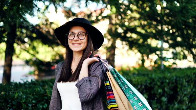 Porträt-der-gut-aussehende-Mädchen-mit-Hut-und-Brille-hält-Einkaufstaschen-Blick-in-die-Kamera-und-lächelnd.-Elegante-Menschen,-Einkauf-und-Jugend-Konzept.