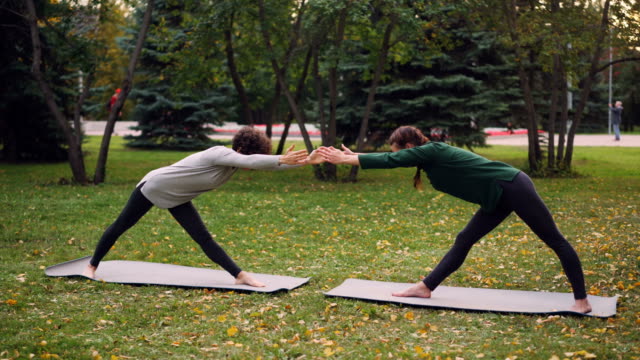 Slim-young-ladies-are-training-outdoors-in-park-doing-hatha-yoga-together-during-pair-practice-and-breathing-fresh-air.-Beautiful-autumn-nature-and-people-are-visible.