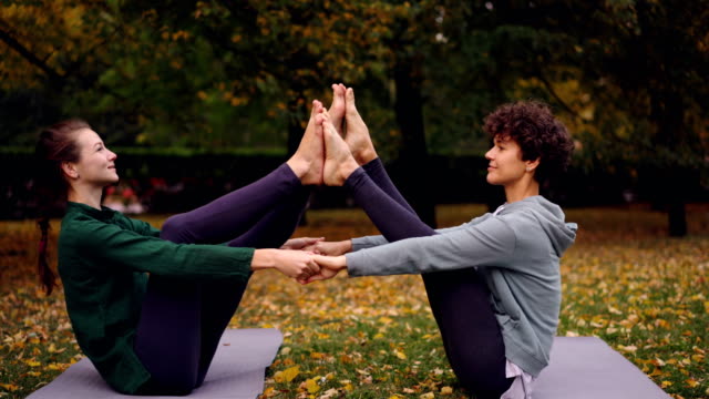 Chicas-atractivas-están-haciendo-ejercicios-de-yoga-en-pareja,-cogidos-de-la-mano-y-poner-pies-juntos-elevando-las-piernas-sentado-en-la-estera-en-el-parque.-Concepto-de-entrenamiento-y-gente-agradable.