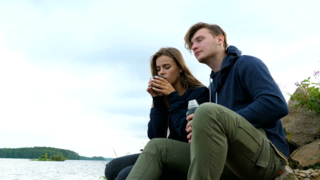 A-guy-with-a-girl-is-sitting-on-the-river-bank-and-drinking-tea-from-a-thermos.