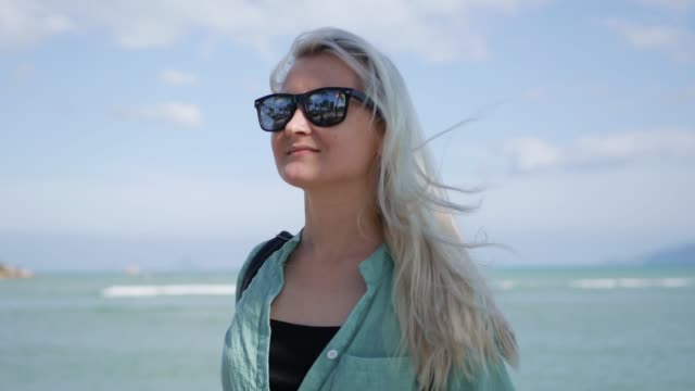 Closeup-of-young-happy-caucasian-woman-with-long-blonde-hair-in-sunglasses-and-green-shirt-standing-and-smiling-near-palm-tree-on-a-blue-sea-background.-Travel-concept