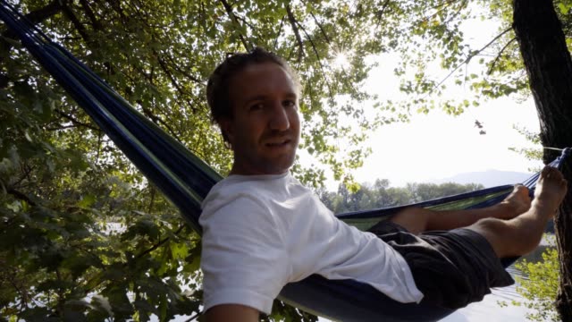 Junger-Mann-nehmen-Selfie-auf-entspannende-Hängematte-am-Fluss-im-Sommer