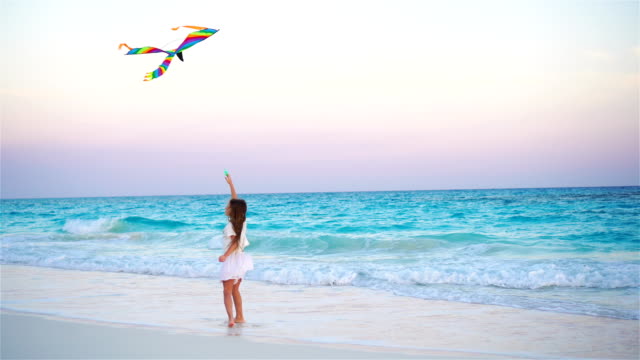 Little-girl-with-flying-kite-on-tropical-beach.-Kid-play-on-ocean-shore.-Child-with-beach-toys-in-slow-motion