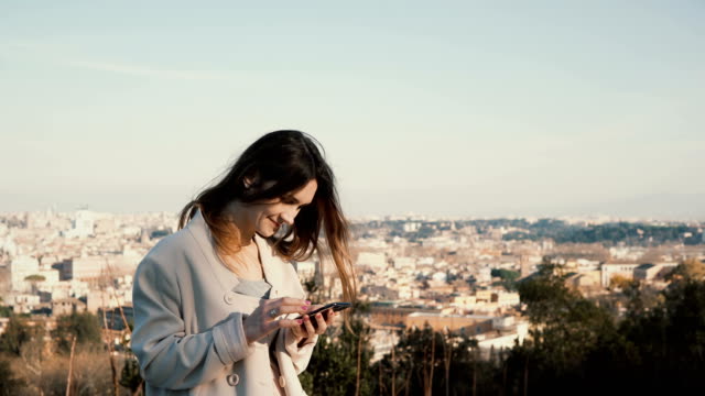 Portrait-of-young-attractive-woman-standing-at-the-panorama-of-Rome,-Italy.-Female-use-the-smartphone-outside