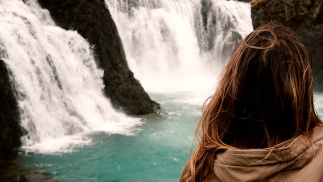 Vista-cercana-de-la-joven-mujer-atractiva-solo-de-pie-y-mirando-en-la-poderosa-cascada-de-Islandia,-onda-de-pelo-en-el-viento