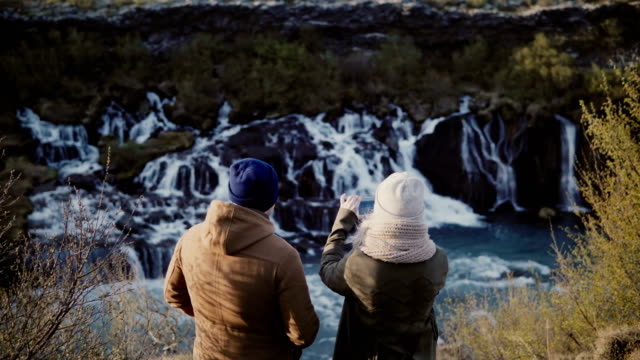 Back-view-of-young-traveling-couple-standing-in-mountains-near-the-waterfalls-in-Iceland-and-taking-photo-on-smartphone