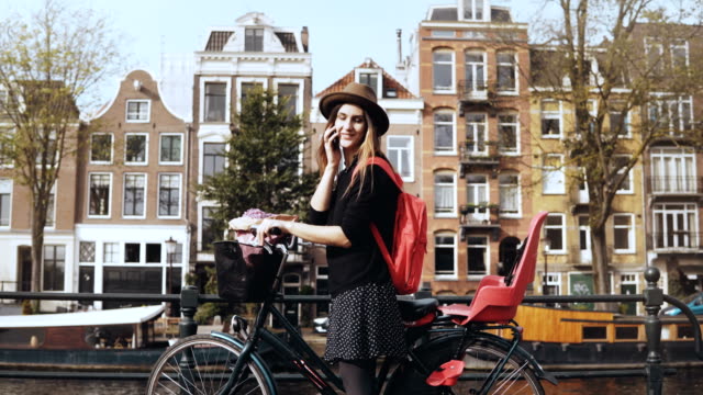 Dama-sonriente-hablando-por-teléfono,-ciclismo-distancia.-Joven-empresaria-en-un-magnífico-puente.-Amsterdam,-Países-Bajos.-4K