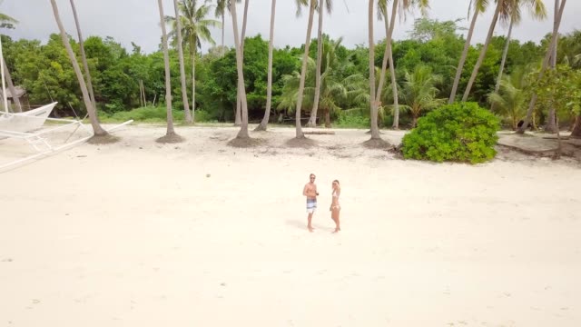 Young-couple-playing-with-drone,-waving-hands-to-the-flying-helicopter.-People-fun-new-technology-vacations-concept.-Shot-on-tropical-beach-in-the-Philippines,-4K-resolution