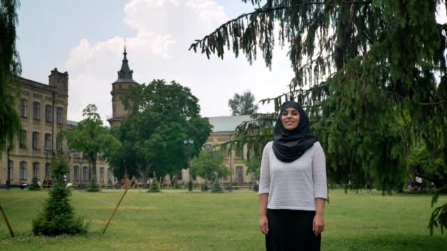 Young-beautiful-muslim-woman-in-hijab-is-standing-and-smiling-in-daytime,-enjoying-weather,-building-on-background,-religiuos-concept