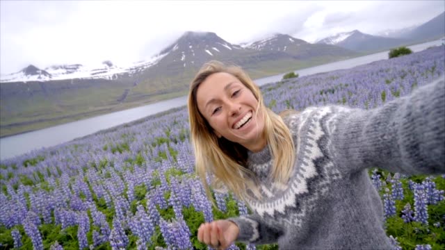 Selfie-portrait-of-tourist-female-in-Iceland-in-the-middle-of-Lupine-purple-flowers,-smiling-hair-in-wind,-wool-seater