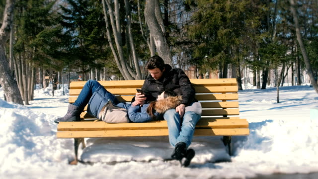 Woman-sleeps,-man-look-at-phone.-Man-and-a-woman-rest-together-on-a-bench-in-the-winter-city-Park.-Sunny-winter-day.