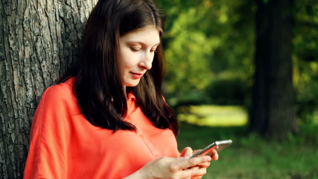 Beautiful-young-lady-is-using-smartphone-resting-in-park-under-tree-and-enjoying-modern-technology-and-summer-nature.-People,-summertime-and-communication-concept.