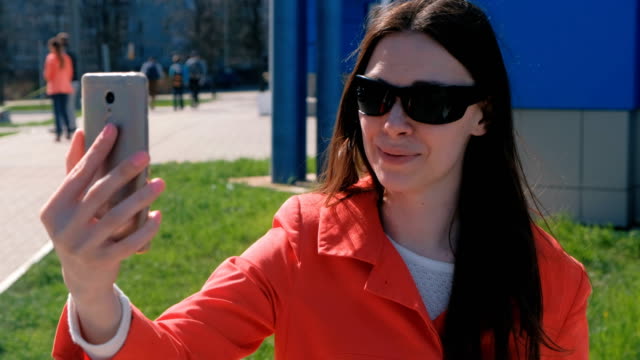 Portrait-of-happy-young-brunette-woman-in-sunglasses-talking-on-video-calling-on-the-phone-beside-blue-building-on-the-street.