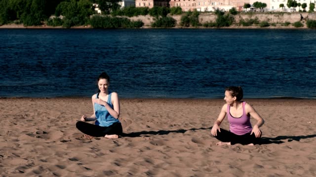 Two-woman-doing-yoga-on-the-beach-by-the-river-in-the-city-and-talking.-Beautiful-view.