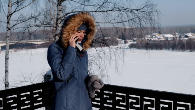 Mujer-en-azul-por-la-chaqueta-con-capucha-de-piel-llamada-en-su-teléfono-celular-en-un-parque-de-invierno.