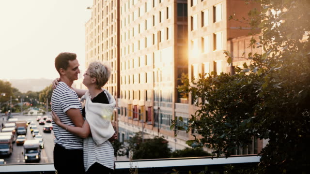 Beautiful-Hispanic-man-and-Caucasian-woman-standing-and-hugging,-holding-hands-on-New-York-City-summer-sunset-bridge