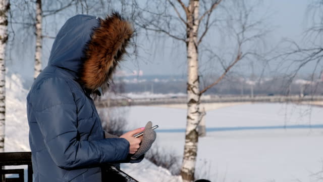 Unrecognizable-woman-in-blue-down-jacket-writes-messaging-in-her-cellphone-in-winter-Park.-Side-view