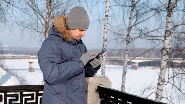 Mann-in-blau-Daunenjacke-mit-Fell-Kapuze-mit-seinem-Handy-für-Web-und-chatten-in-einem-Winter-Park.