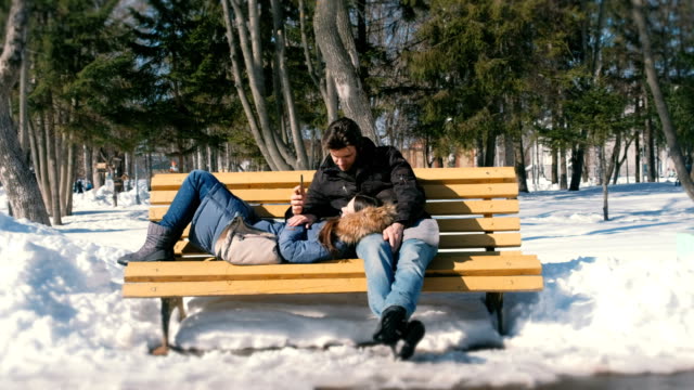 Man-and-a-woman-rest-together-on-a-bench-in-the-winter-city-Park.-Sunny-winter-day.-Look-something-on-mobile-phone.