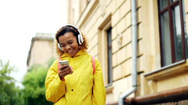 Dolly-shot-of-cheerful-young-lady-listening-to-music-with-headphones,-singing-and-using-smartphone-walking-outdoors-in-modern-city.-Fun,-people-and-technology-concept.