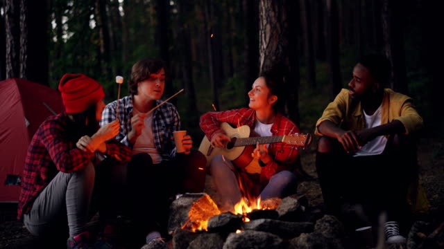 Happy-men-and-women-are-sitting-around-fire-cooking-marshmallow-playing-the-guitar-and-singing-during-hike-in-forest.-Young-people-are-having-fun-and-enjoying-nature.