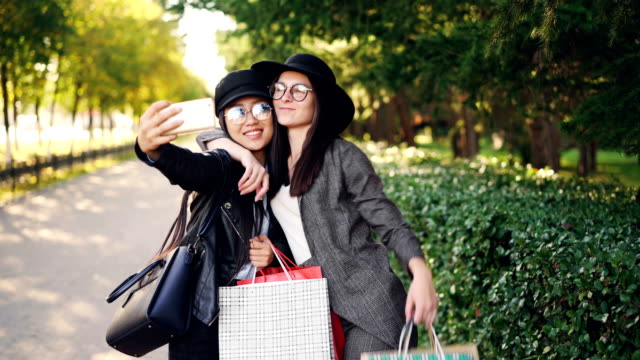 Excited-female-friends-are-taking-selfie-using-smartphone-and-holding-shopping-bags-with-purchases-on-sunny-autumn-day.-Modern-technology-and-youth-concept.