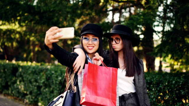 Attraktive-Asiatin-nimmt-Selfie-mit-Freundin-kaukasischen-nach-Tag-Holding-Einkaufstaschen-und-mit-Smartphone.-Moderner-Lifestyle,-Technik-und-Jugend-Konzept.