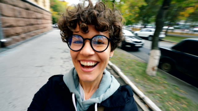 Point-of-view-shot-of-carefree-young-lady-with-short-hair-and-glasses-looking-at-camera-and-laughing-turning-around-in-the-street-having-fun-on-autumn-day.