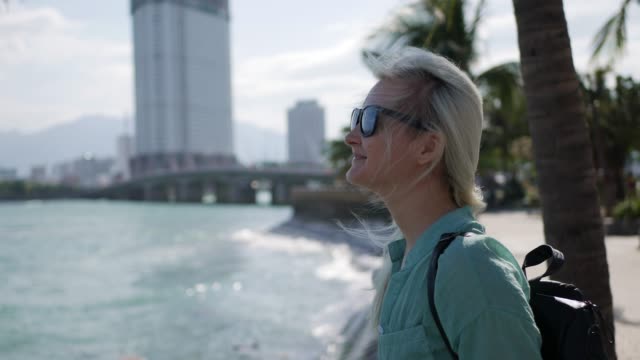 Young-happy-caucasian-woman-with-long-blonde-hair-in-sunglasses-and-green-shirt-standing-and-smiling-near-palm-tree-on-a-park-and-sea-background.-Travel-concept