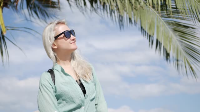 Young-happy-caucasian-woman-with-long-blonde-hair-in-sunglasses-and-green-shirt-standing-and-smiling-near-palm-tree-on-a-blue-sky-background.-Travel-concept