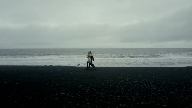 Aerial-view-of-young-couple-walking-on-black-volcanic-beach.-Man-and-woman-have-a-date-on-shore-of-the-sea-in-Iceland