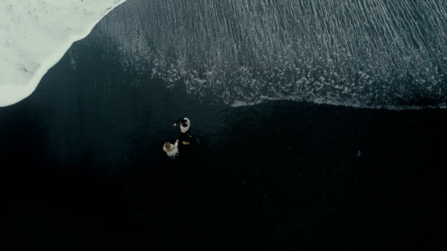 Top-Luftaufnahme-des-jungen-Paares-am-Ufer-des-Meeres-in-schwarzen-Strand-in-Island-stehen-und-das-Wasser-genießen