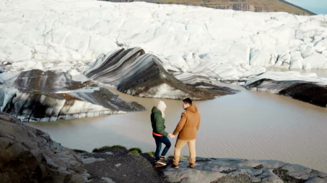 Young-beautiful-couple-standing-on-the-shore-of-the-lake-in-Vatnajokull-ice-lagoon-in-Iceland-and-looking-on-glaciers