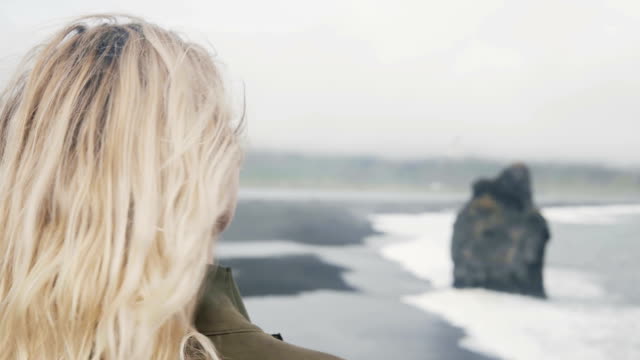 Joven-rubia-de-pie-en-la-orilla-del-mar-y-mirando-en-los-dedos-del-Troll-de-las-rocas-de-interés-en-Islandia-en-día-nublado