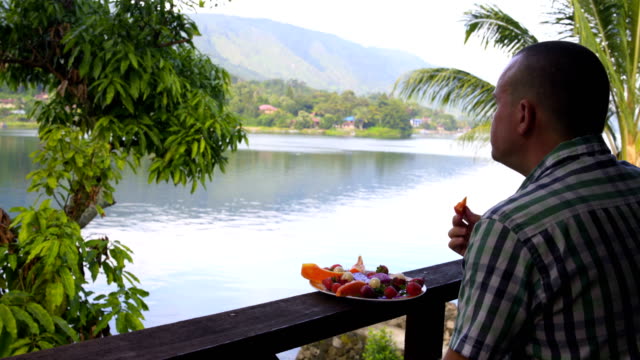 Un-hombre-está-comiendo-fruta-y-mirando-el-lago-y-las-montañas