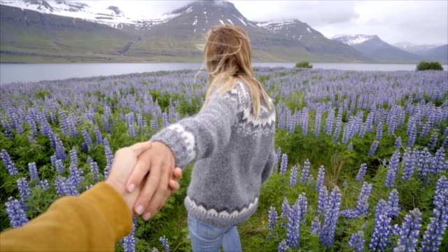Follow-me-to-Iceland,-girlfriend-leading-man-to-flower-lupine-field-near-lake-and-mountains-People-travel-concept--4K-video