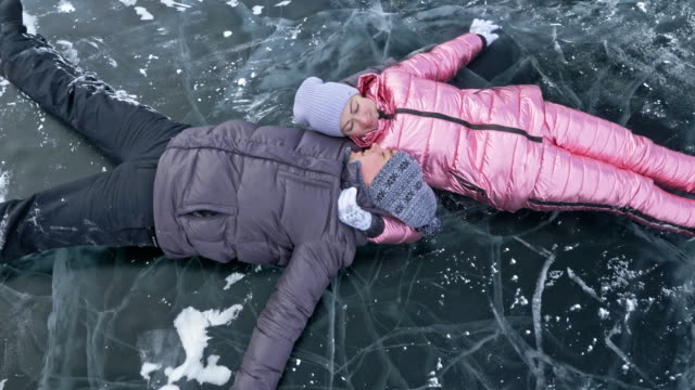 Young-couple-has-fun-during-winter-walk-against-background-of-ice-of-frozen-lake.-Lovers-lie-on-clear-ice-with-cracks,-have-fun,-kiss-and-hug.-View-from-above.-Honeymoon.-Love-story.