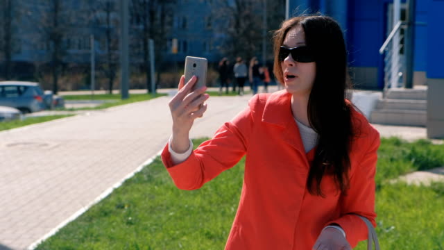 Young-brunette-woman-in-sunglasses-talking-on-video-calling-on-the-phone-beside-blue-building-on-the-street.