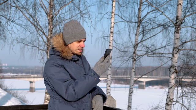 Mann-in-blau-Daunenjacke-mit-Fell-Kapuze-mit-Handy-für-Video-Chat-und-lächelnd-in-Winter-Park.