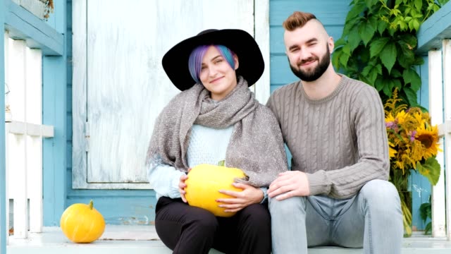 Young-woman-and-man,-family,-sitting-on-porch-of-village-house-with-pumpkins.