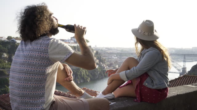 Young-smiling-lady-and-man-drinking-from-bottle-sitting-on-roof