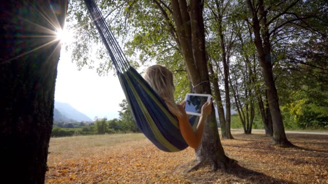 Young-woman-on-hammock-at-sunset-reading-on-digital-tablet-using-mobile-apps-on-wireless-technology.-People-travel-modern-life-concept