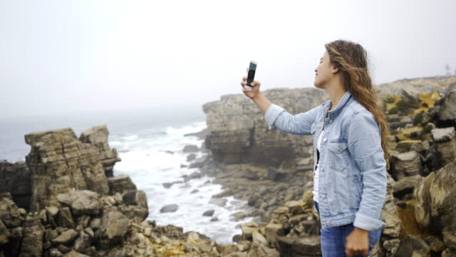 Young-woman-taking-selfie-on-smartphone-on-stone-shore-near-water