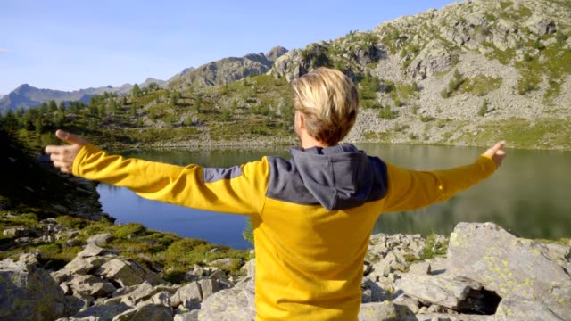 Young-man-hiker-on-trail-in-Summer-by-stunning-alpine-lake-arms-outstretched-celebrating-personal-goal.-Young-man-standing-in-nature-arms-wide-open
