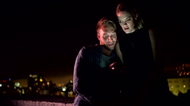 Young-couple-looking-at-smart-phone-on-rooftop-at-night