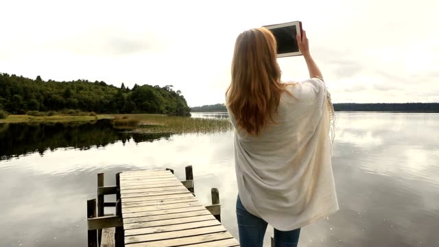 Young-adult-relaxes-on-jetty-above-lake,-uses-digital-tablet